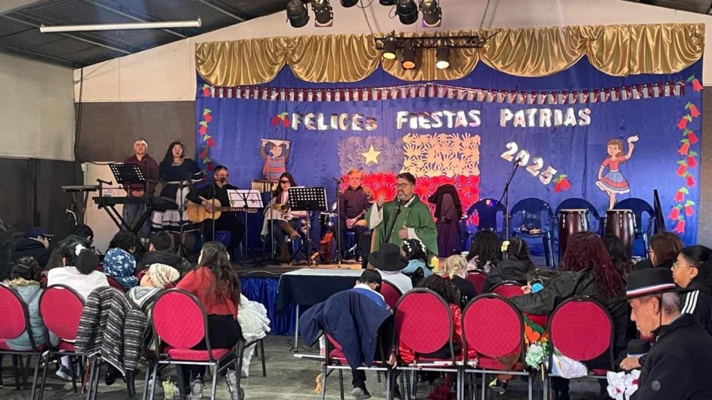 Imagen lejana donde se ve al cura, el escenario con la banda atrás y algunos de los asistentes sentados