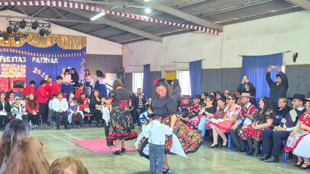 Vista general de la fiesta del 18 en el medio bailando cueca dos pequeños estudiantes de Colegio Santa Lucía junto a sus profesoras. Se ve también a los asistentes sentados mirando el baile.