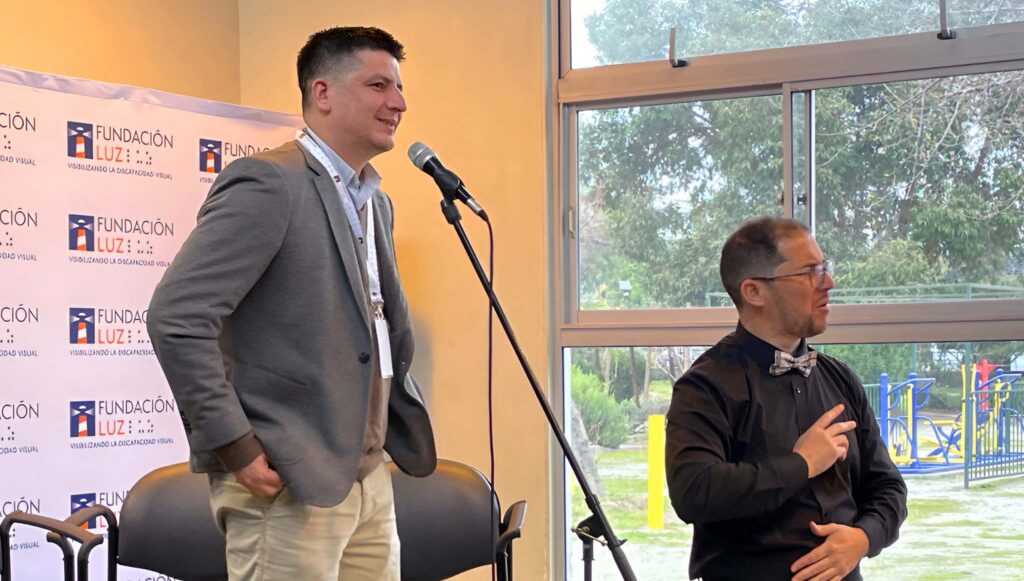 Fotografía tomada en un evento de Fundación Luz. En primer plano, aparece Álvaro Venegas, de pie junto a un micrófono, vistiendo chaqueta gris, camisa celeste y pantalón beige, mientras sonríe al público. A su lado, un intérprete de lengua de señas, con camisa negra y corbatín gris, traduce su mensaje. Al fondo se observa el logotipo de Fundación Luz y una gran ventana con vista a un área verde exterior.