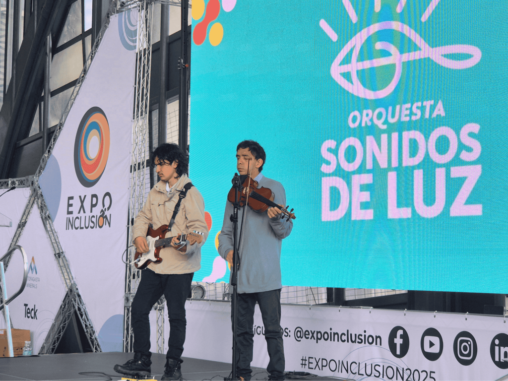 Dos integrantes de la Orquesta Sonidos de Luz, guitarra eléctrica y violinista, tocando sobre el escenario de Expo Inclusión.