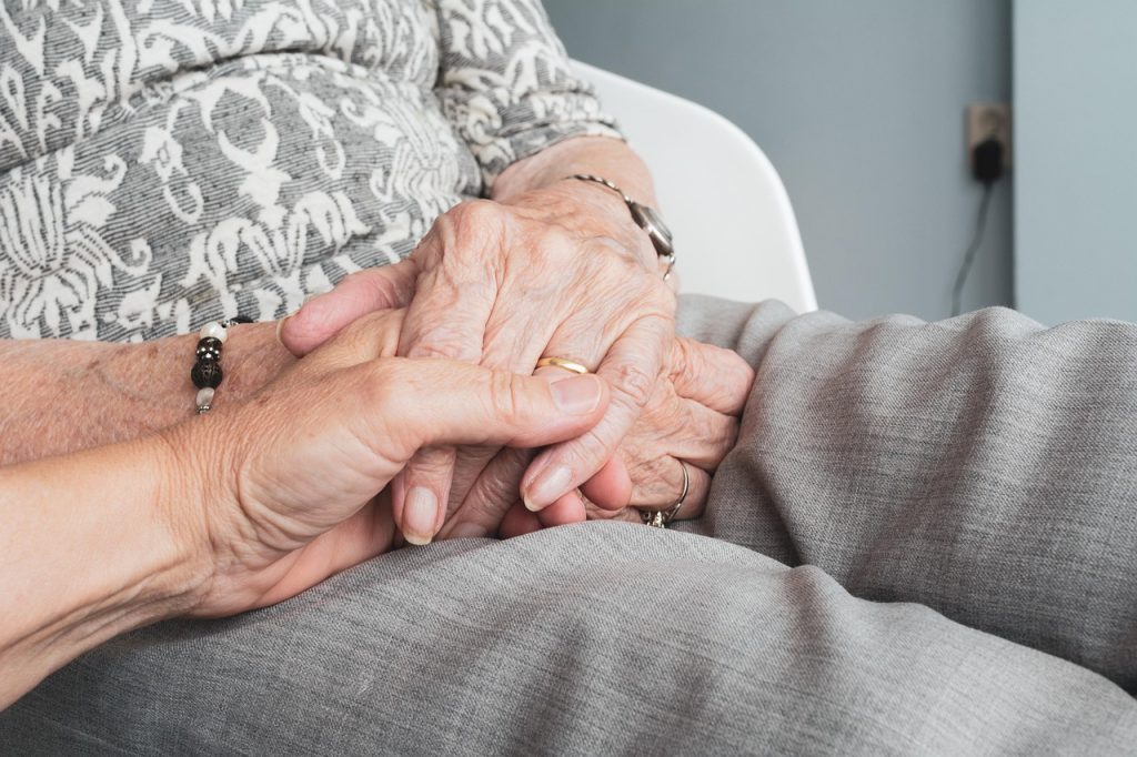Mano de una mujer de la tercera edad sostenida por la mano de un hombre m&aacute;s joven. Ambas est&aacute;n apoyadas en el regazo de la mujer mayor.