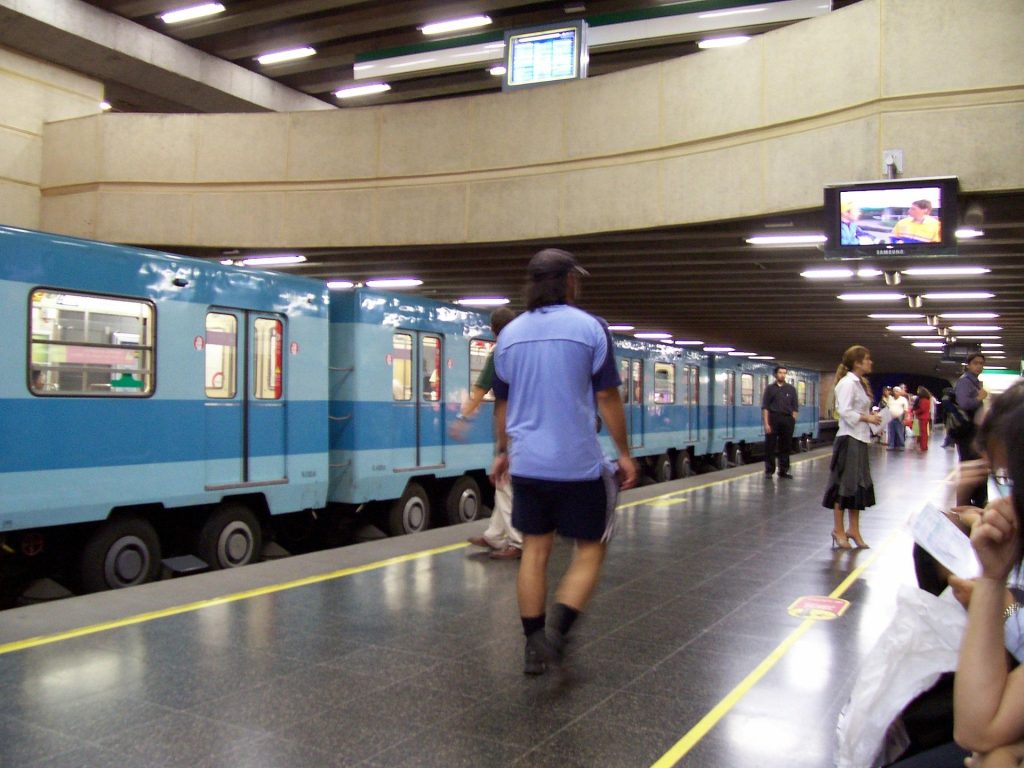 Andén de una estación del metro de Santiago. Algunas personas caminan, otras esperan sentadas. En la dirección contraria, está detenido un tren mientras suben pasajeros.