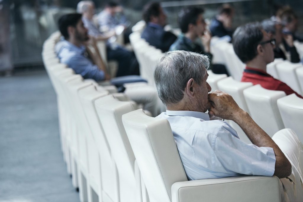 Foto desde el costado de personas sentadas mirando hacia adelante y escuchando con atenci&oacute;n.